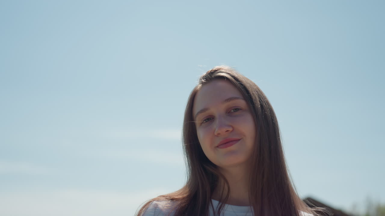 Close up of young woman with straight brown hair and soft smile standing outdoors on sunny day, blue sky and blurred pine tree and building roof in background