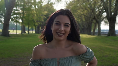 slow motion portrait of a gorgeous hispanic latino young woman looking at the camera and show emotions from serious to smiling and laughing with a beautiful british park in the background