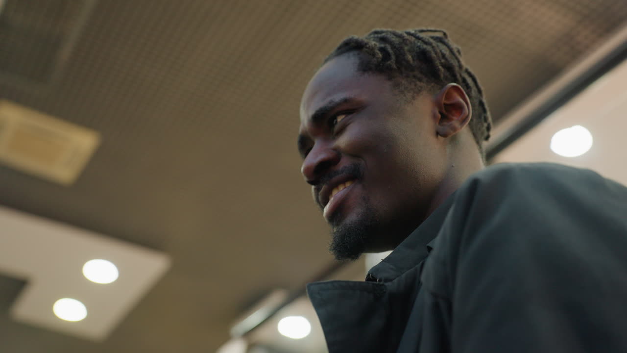 Close up of young man with braided hair smiling warmly while interacting with someone on ascending escalator inside building with ceiling lights and mesh panels