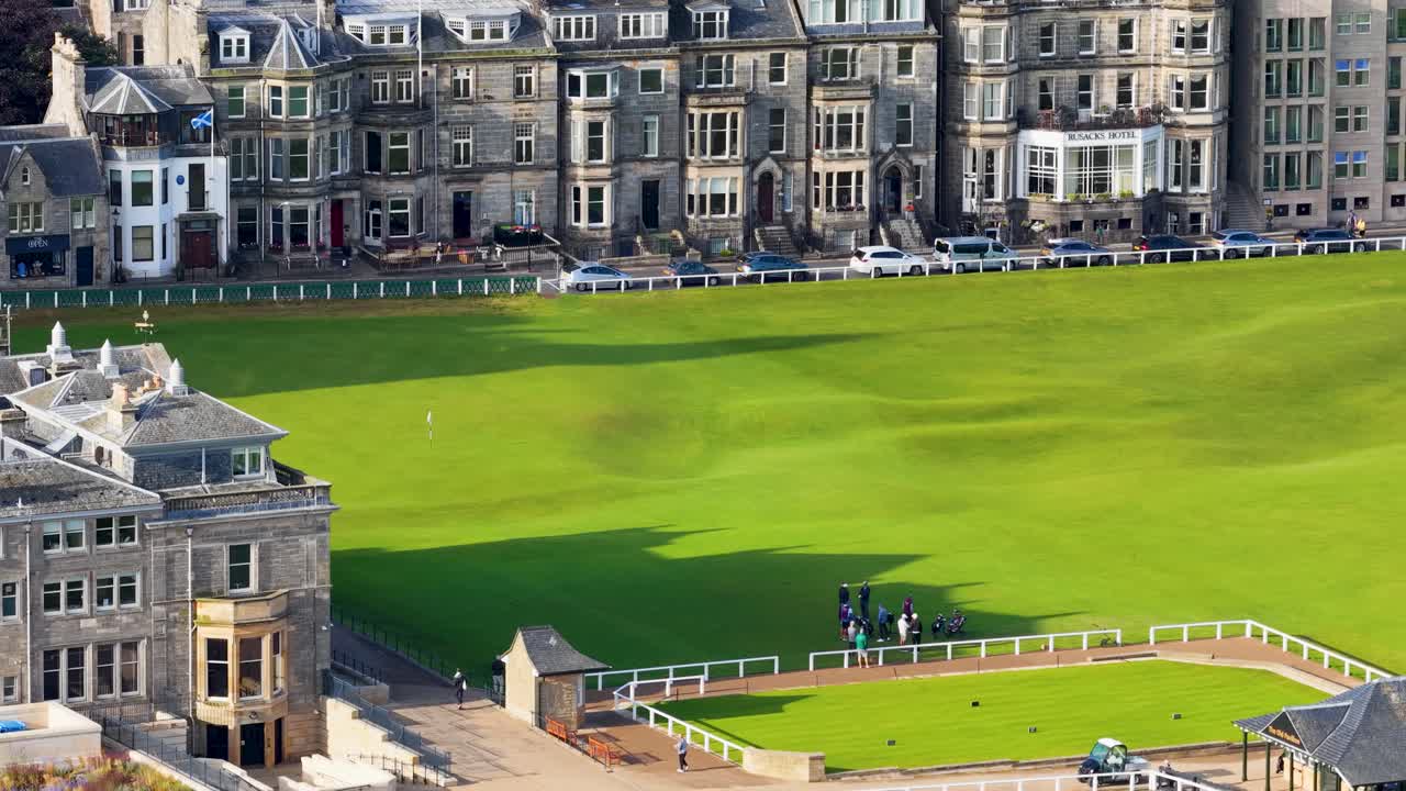 Drone captures golfers walking near iconic clubhouse on sunlit, lush green fairway in Scotland
