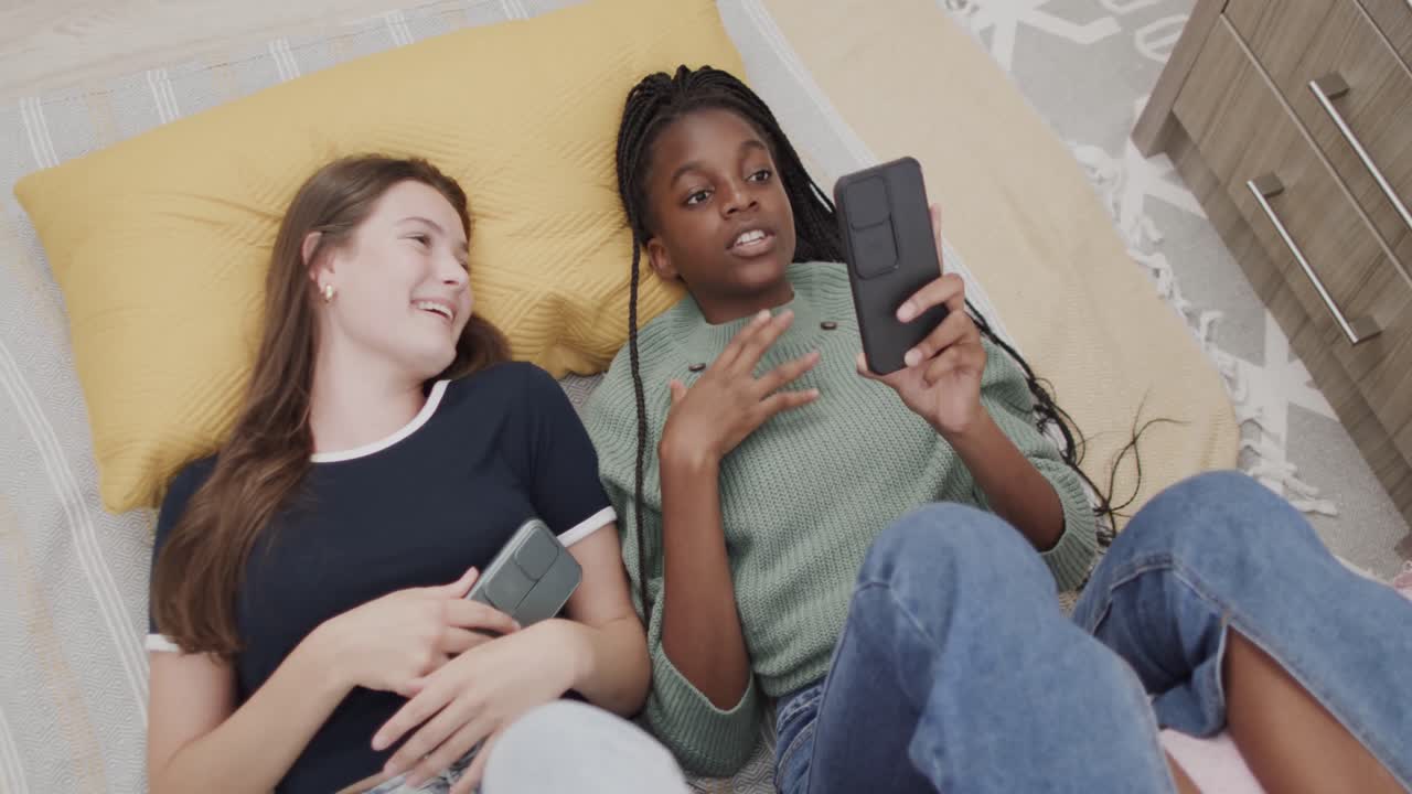 Happy diverse teenage female friends lying on bed using smartphones and laughing