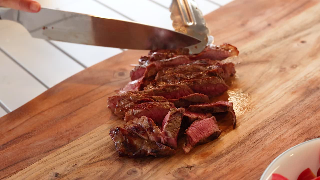 A person slices a cooked beef steak on a wooden board in a well-lit kitchen setting
