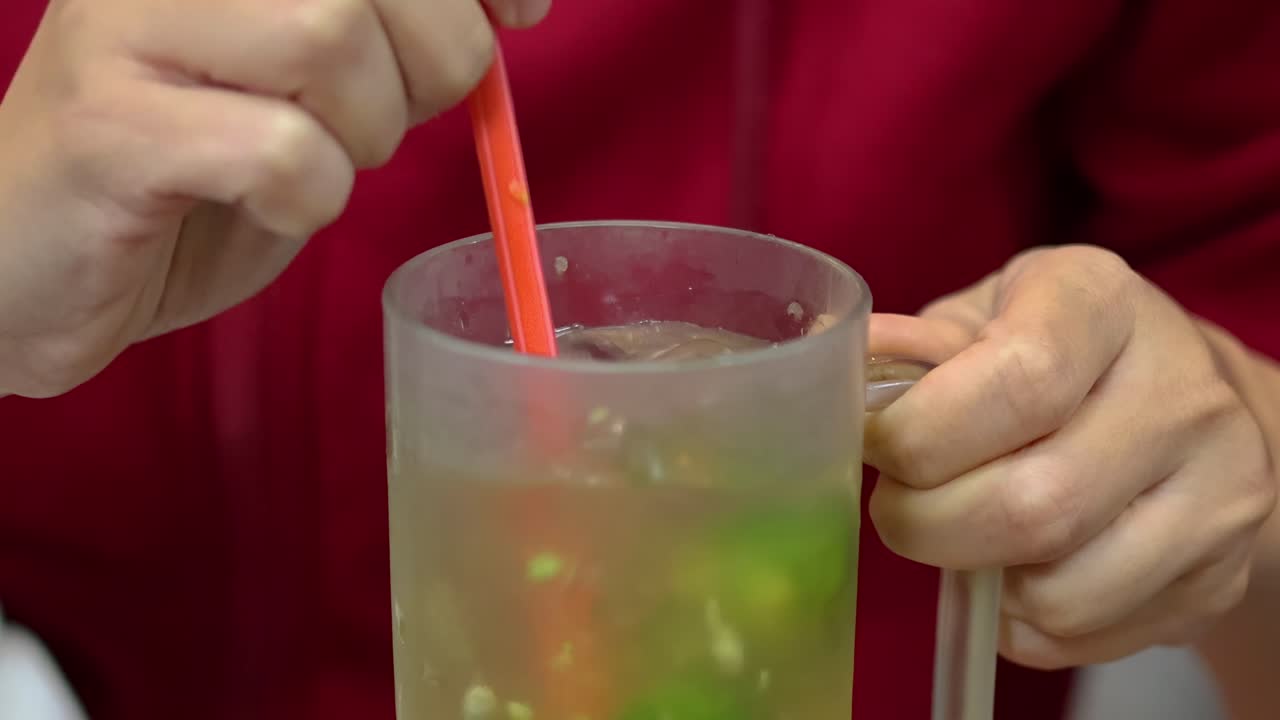 Hands stirring a lime and ice-filled beverage with a red straw in a frosted mug.