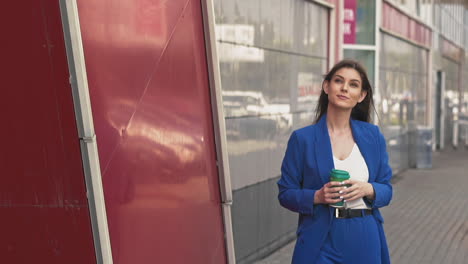 woman in blue suit walks holding cup of coffee along street