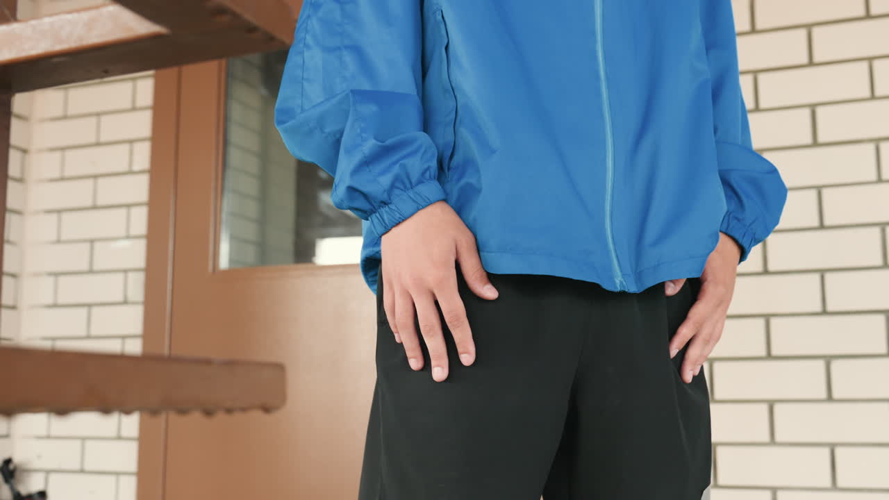 階段のそばに立ち、ポケットに手を入れ、黒いパンツをはき、レンガの壁を背景にした、青いジャケットを着たアスリート、雨の雰囲気、都会の脇道での休憩、選手が練習の合間に待つ間のかすかな動き、クローズアップフレーミング。