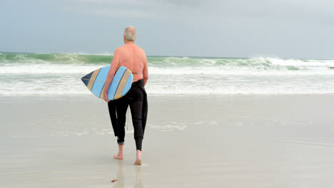 Rückansicht-Eines-Alten-Kaukasischen-Mannes,-Der-Mit-Einem-Surfbrett-Am-Strand-Spaziert,-4k