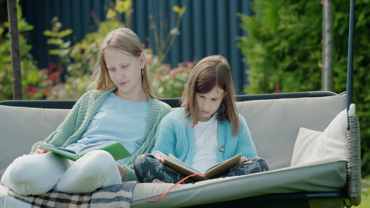 Due ragazze leggono libri, sedute su un'altalena da giardino nel cortile di casa.