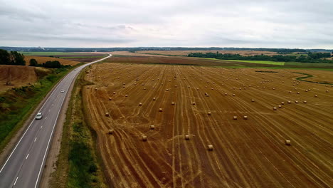 Farmland-with-rolls-of-baled-hay-near-a-country-road---aerial-flyover
