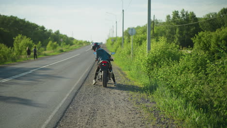 una mujer en una motocicleta mirando hacia atrás mientras comienza su bicicleta por la carretera, un arbusto verde exuberante bordea la carretera a medida que los coches se acercan desde la dirección opuesta, y la gente camina a lo largo de la carretera