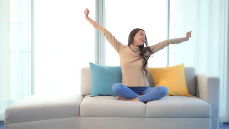 asian woman stretching her arms sitting on sofa at home near window