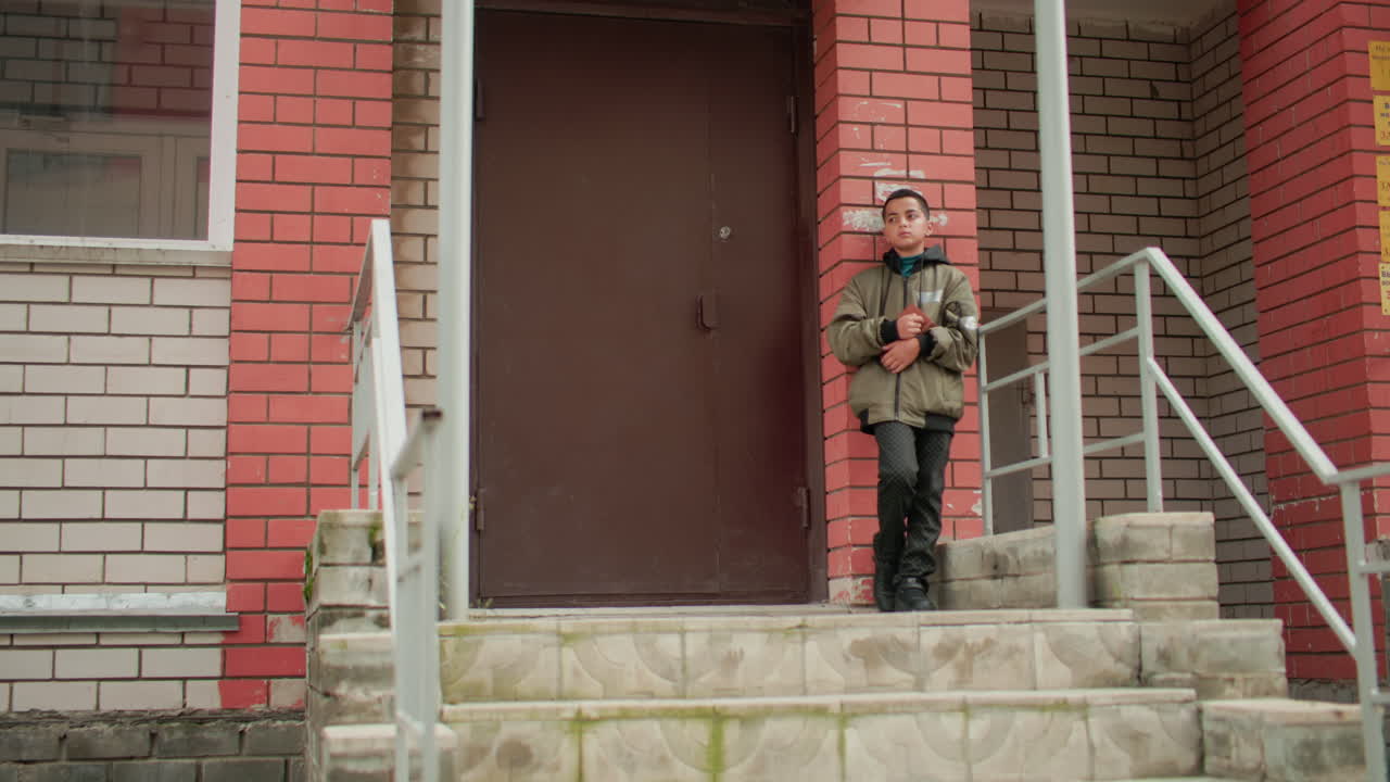 Adolescent in hooded jacket leans on brick wall near building entrance holding brown book, looking thoughtfully ahead while standing on outdoor concrete staircase with metal railings