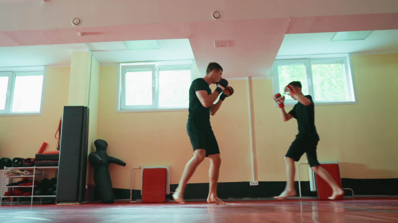 Two wrestlers sparring in gym on red mat, facing each other in combat stance, wearing gloves, barefoot, practicing martial arts with focus on strength, endurance, discipline, fighting technique