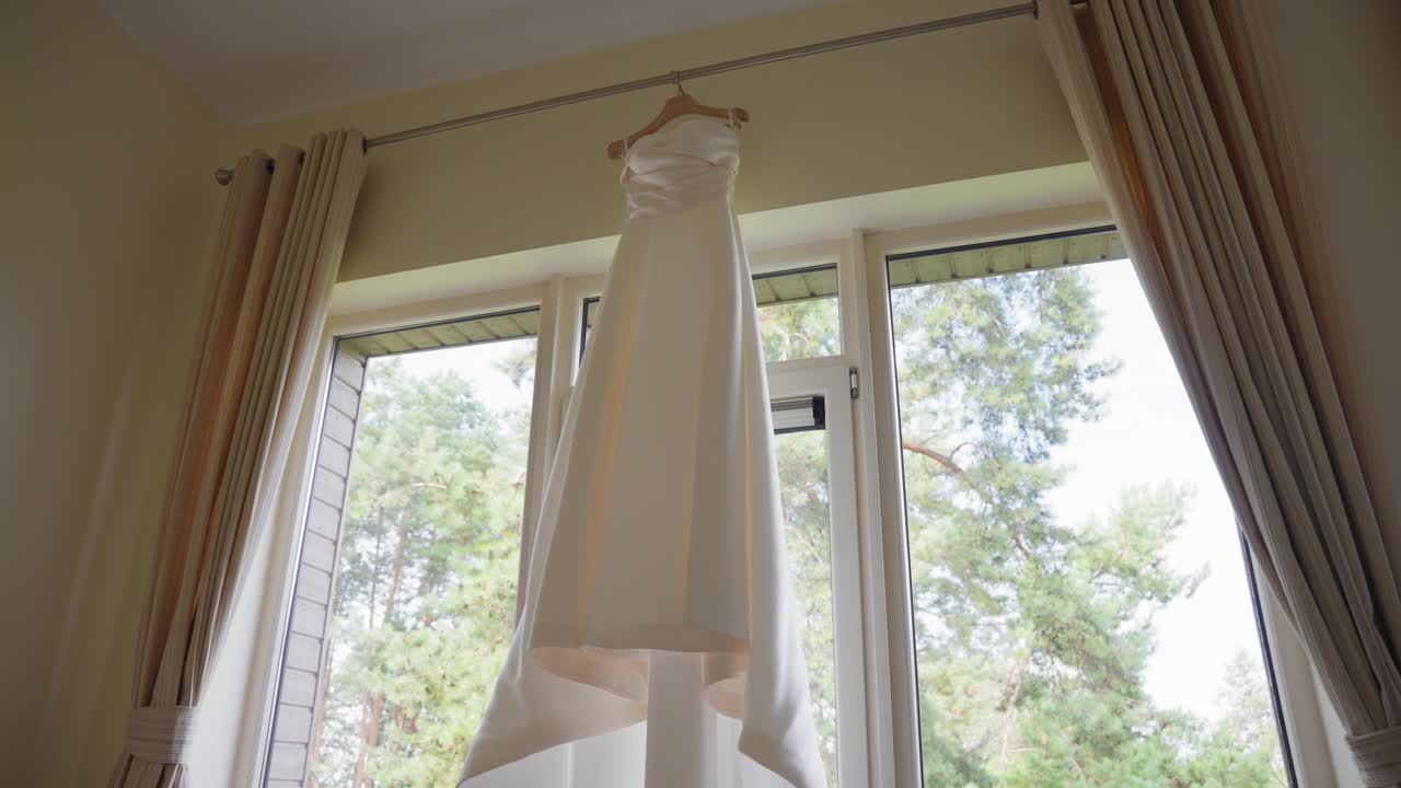 A stunning white wedding dress elegantly hangs by a sunlit window. The soft natural light streams through the trees outside, creating a serene and romantic ambiance