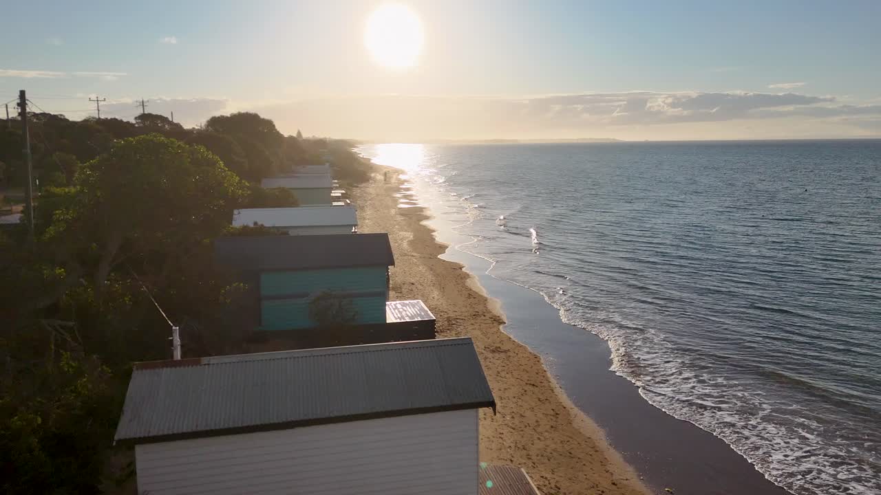 Drone glides above colorful beach huts at sunrise, capturing tranquil shoreline and golden morning light