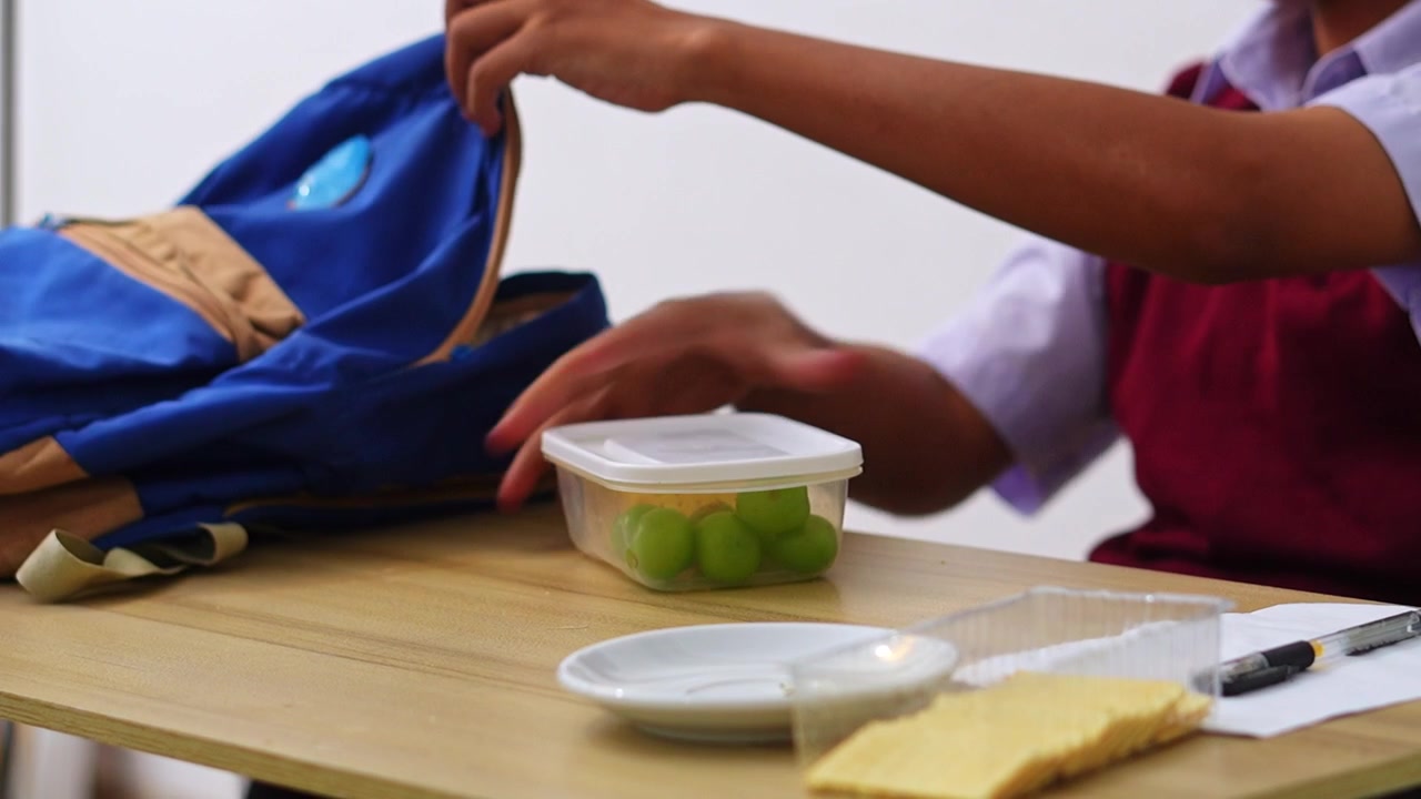 Estudiante con chaleco granate guardando una fiambrera de plástico llena de uvas y galletas saladas en la mochila