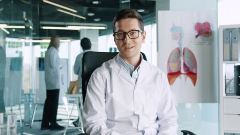 Caucasian-male-doctor-in-glasses-sitting-in-clinic-at-workplace-looking-at-camera-and-speaking-on-medical-consultation