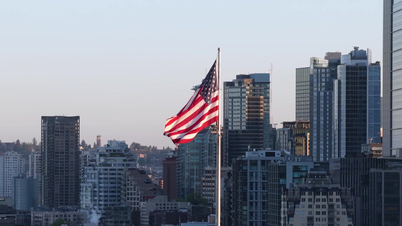 Bandiera americana sventola davanti allo skyline di Seattle con lo Space Needle e imponenti grattacieli di vetro del centro che brillano sotto una morbida luce serale rosa a Seattle, Washington, USA. Veduta aerea con drone.