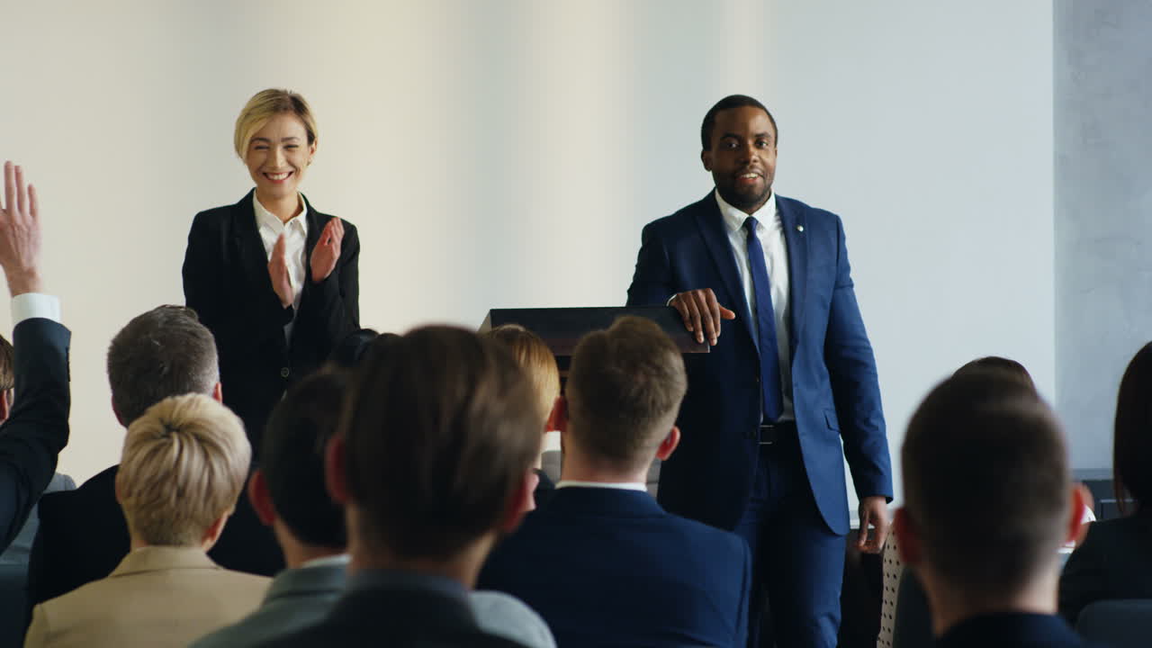Young Caucasian businesswoman standing on a podium and presenting her colleague African American man who is sitting on a chair and coming to the stage
