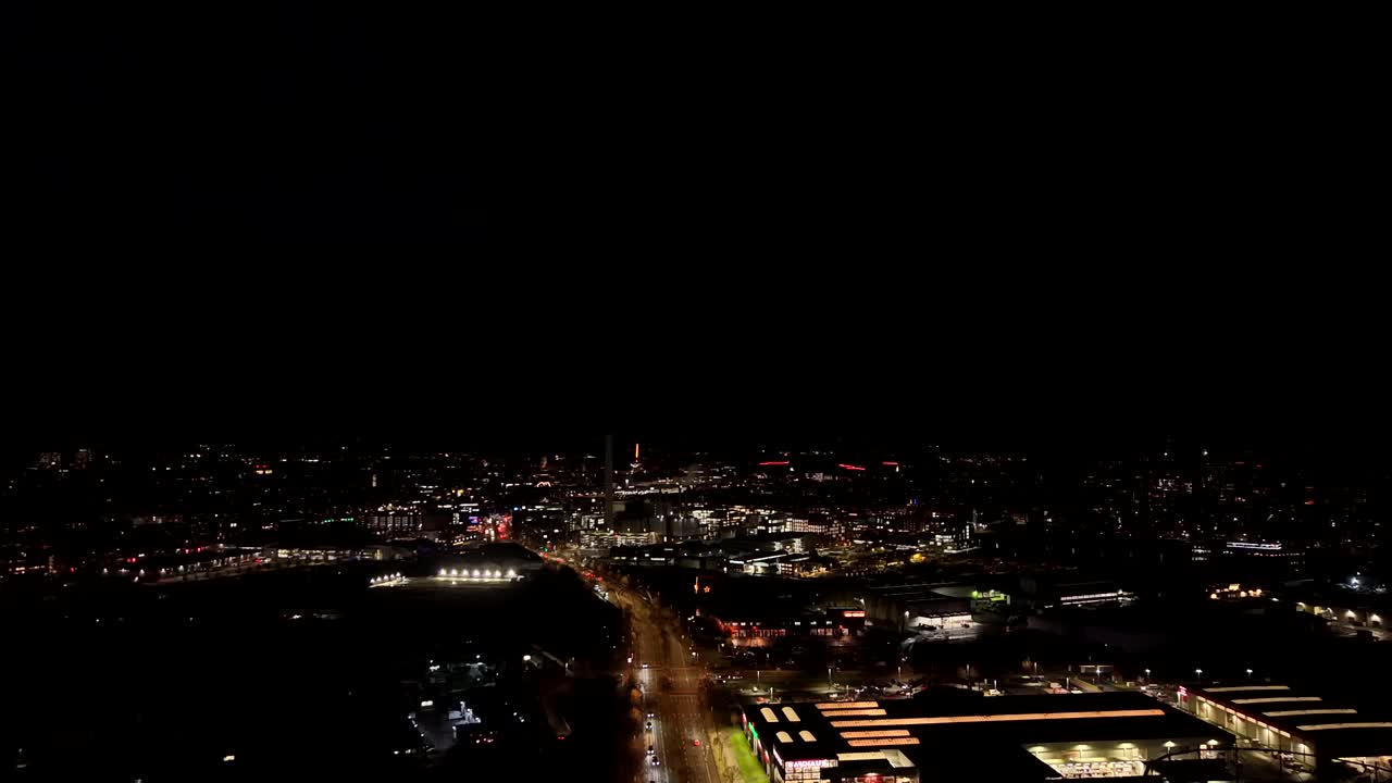 Aerial wide shot over American city at night showing glowing warehouses and bright lantern lights along busy road. Urban skyline stretches across dark landscape in illuminated United States town