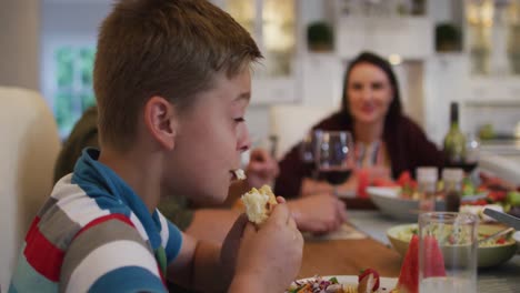 Smiling-caucasian-son-eating-at-table-during-family-meal,-parents-in-background