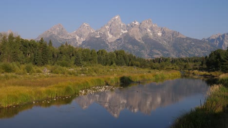 A pristine river winds through lush greenery beneath snow-capped peaks. Majestic mountains reflect in calm waters, capturing the serene beauty of alpine wilderness in a tranquil summer scene