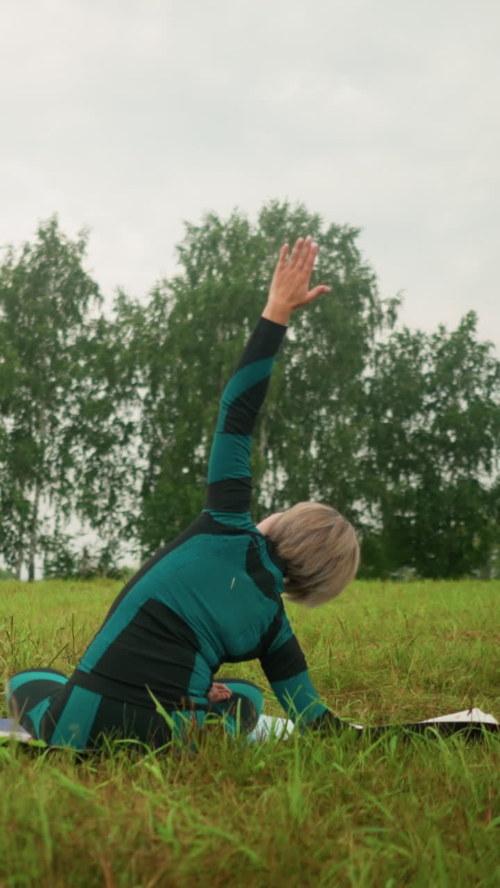 vista trasera de la mujer acostada en un lado de la alfombra de yoga, practicando la postura de flexión lateral con el brazo extendido, sentada en un vasto campo de hierba bajo un cielo nublado, rodeada de naturaleza y árboles en la distancia