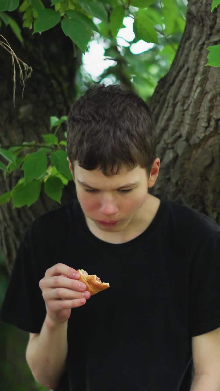 hermanos disfrutan de un descanso mientras comen bocadillos bajo un árbol en un bosque pacífico, uno de los niños se apoya en el árbol mientras el otro está de pie, ambos rodeados de exuberante vegetación