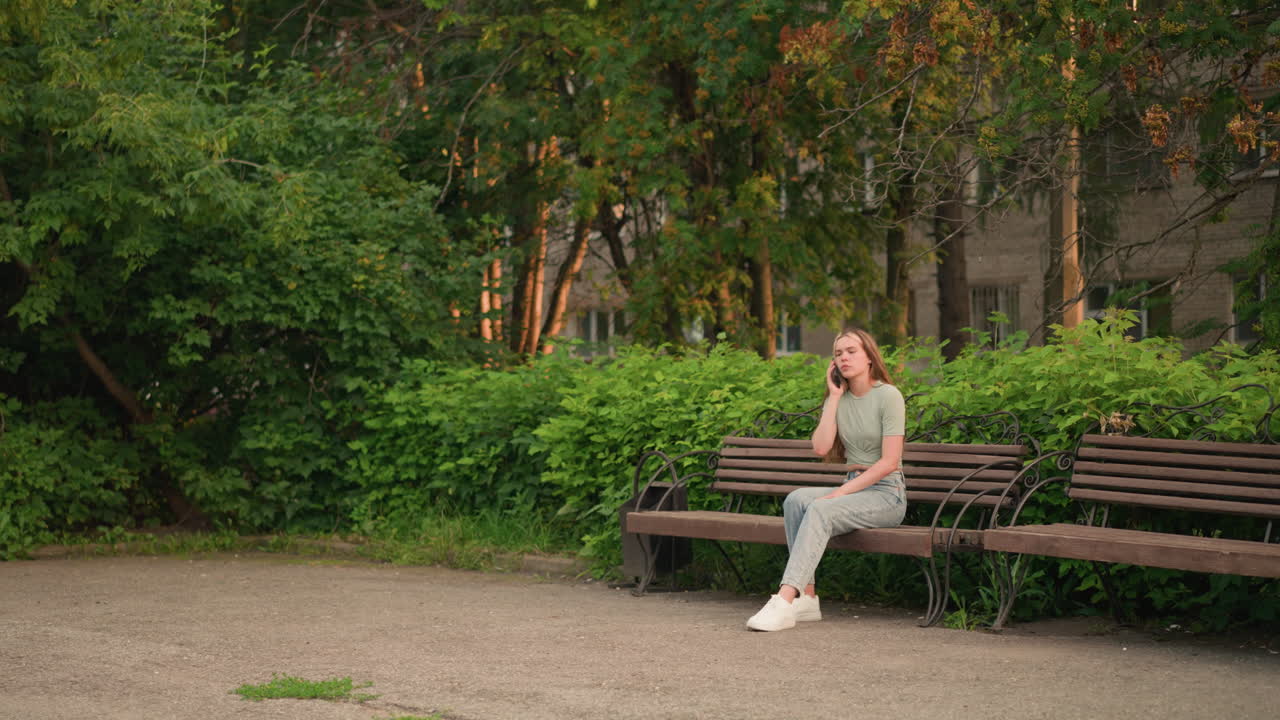 전화 통화 중인 여인은 나무 벤치에 앉아서, 잎자루를 통해 어내는 은 초록색과 나무, 그리고 뒷면에 있는 건물에 둘러싸여 생각 깊게 내려다보고 있습니다.