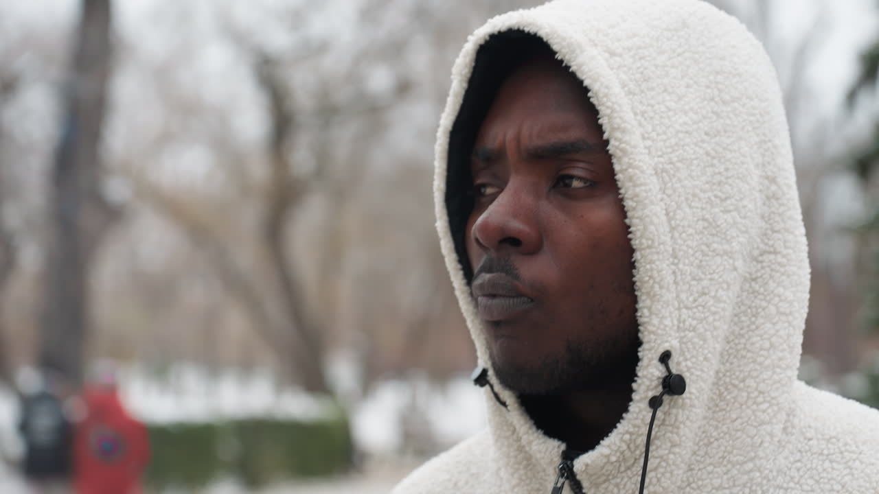 серьезный молодой человек в белом капюшоне ест во время прогулки, глубоко в мыслях с размытым зимним фоном голых деревьев, добавляя капризную и интроспективную атмосферу в сцену городского парка