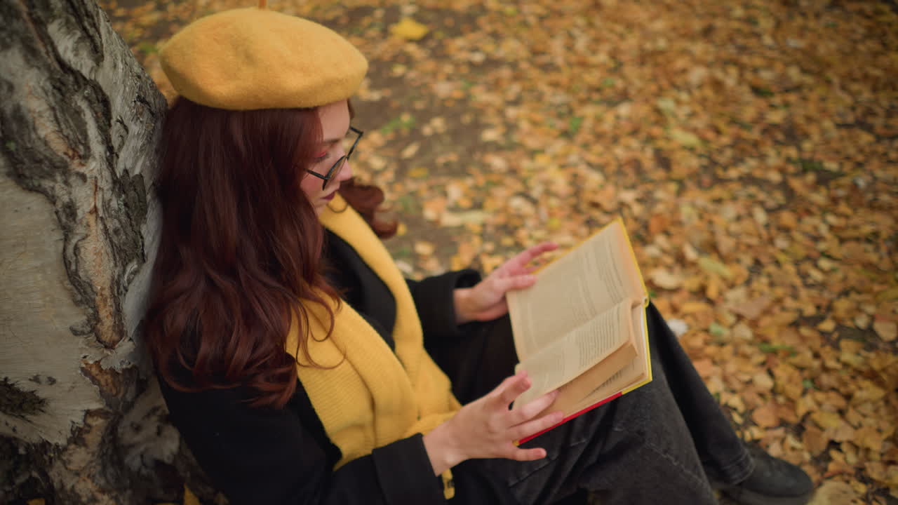 vista aérea de una mujer con el cabello rojo ondulado con boina amarilla y gafas de sol hojeando un libro en un entorno pacífico de otoño, rodeada de hojas caídas, inmersa en el pensamiento y la literatura