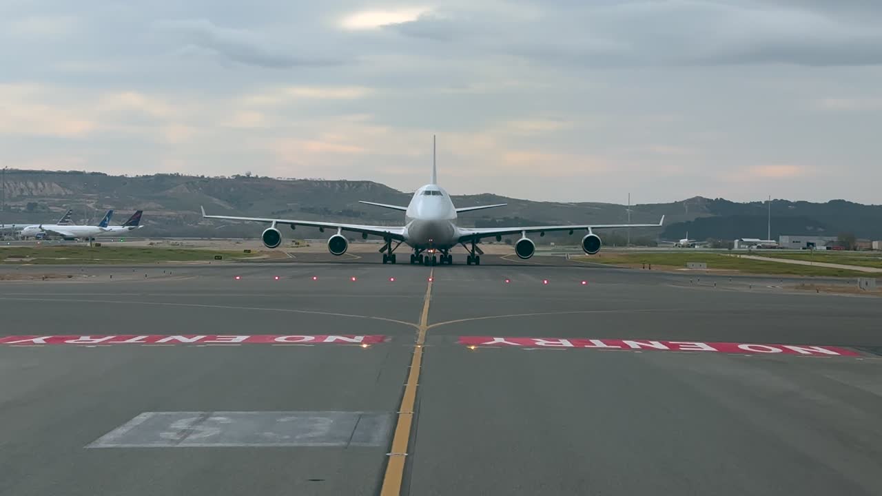 Vista frontale esterna di un enorme jet a fusoliera larga a quattro motori che rulla lentamente attraverso la via di rullaggio dell'aeroporto di Madrid di prima mattina. Scena catturata dall'abitacolo di un altro jet che rulla in direzione opposta