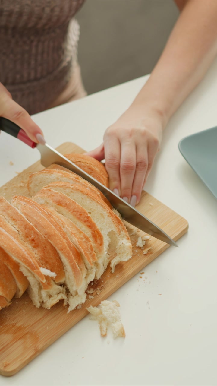 Woman slices loaf of bread on wooden cutting board with steady hand movements. fresh golden brown crust and soft interior are visible as knife cuts through evenly. light blue plate sits