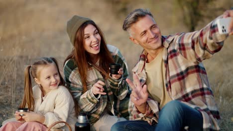 A-happy-middle-aged-man,-together-with-his-wife,-a-brunette-girl-in-a-Green-checkered-shirt,-and-a-little-daughter,-take-a-selfie-using-a-white-phone-during-their-picnic-and-vacation-outside-the-city-in-the-summer