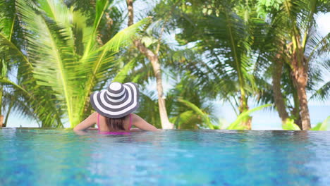 lonely woman in infinity swimming pool on tropical destination, back view of female in floppy hat looking at exotic vegetation, full frame