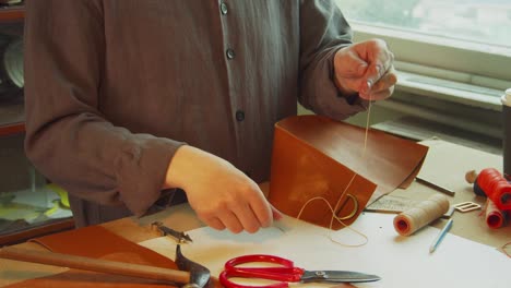 a young, experienced tailor's apprentice threads a needle for sewing a genuine leather product in a leather workshop