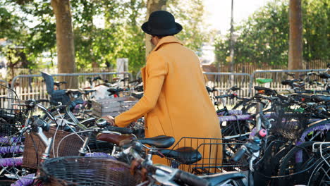 young woman wearing unbuttoned yellow pea coat, black hat and sunglasses parking her bicycle, putting on crossbody bag and walking away smiling, side view, tracking shot