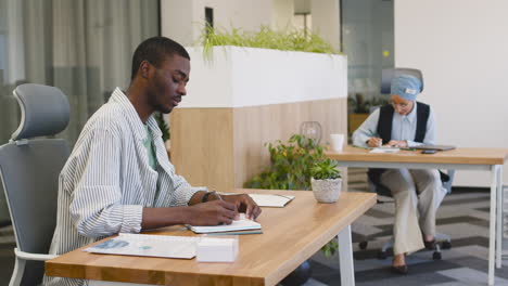 Joven-Trabajador-Sonriente-Trabajando-Tomando-Notas-Sentado-En-Su-Escritorio