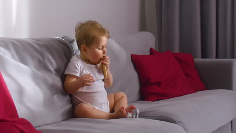 front view of a baby sitting on sofa in living room at home while bitting a wooden bracelet