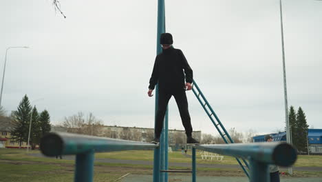 a young boy dressed in a black outfit and glasses walks carefully on an iron rail while another boy in a gray sweater rests his right hand on an adjacent iron structure