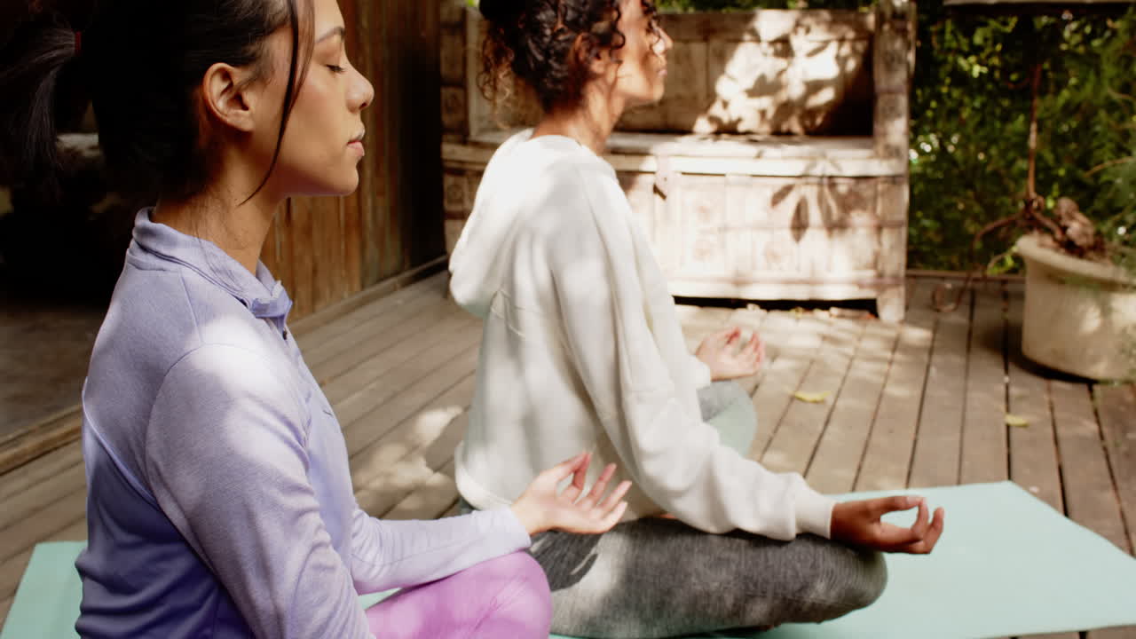 Two multiracial female friends meditating on porch on wooden deck, enjoying peaceful moment