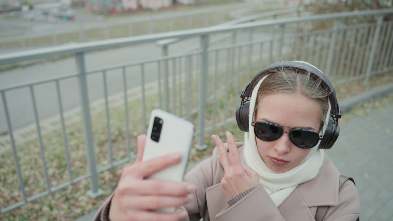 Una giovane donna si fa un selfie su una passerella cittadina, con cuffie e occhiali da sole, avvolta in sciarpa e cappotto. Gesticola alla telecamera per un vlog, mostrando un'espressione giocosa e sicura, con ringhiera metallica e strada sullo sfondo.