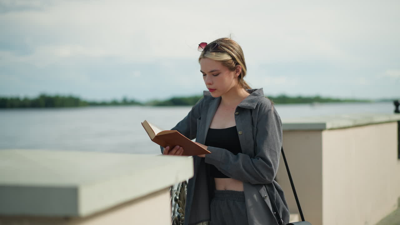 Woman in grey clothing leans her right hand on an iron fence while reading a book by the riverfront, the wind blows gently, with trees and the water visible in the background