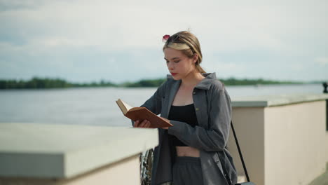 woman in grey clothing leans her right hand on an iron fence while reading a book by the riverfront, the wind blows gently, with trees and the water visible in the background