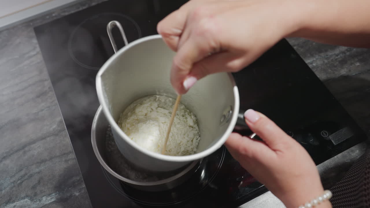 Female chandler melts tallow flakes inside metal mixing bowl placed in pot of hot water on electric stove, using double boiler method to safely liquefy wax substance for candle making in home kitchen