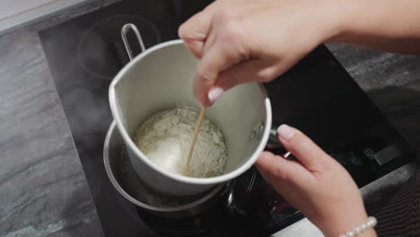 female chandler melts tallow flakes inside metal mixing bowl placed in pot of hot water on electric stove, using double boiler method to safely liquefy wax substance for candle making in home kitchen
