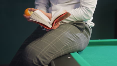 lower body of person wearing grey trousers sitting on green pool table while holding book and billiard ball. casual posture with relaxed atmosphere, emphasizing focus and leisure pool hall setting