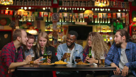 a company of young men and women sitting in a bar at a table drinking beer and laughing telling each other funny gossip.