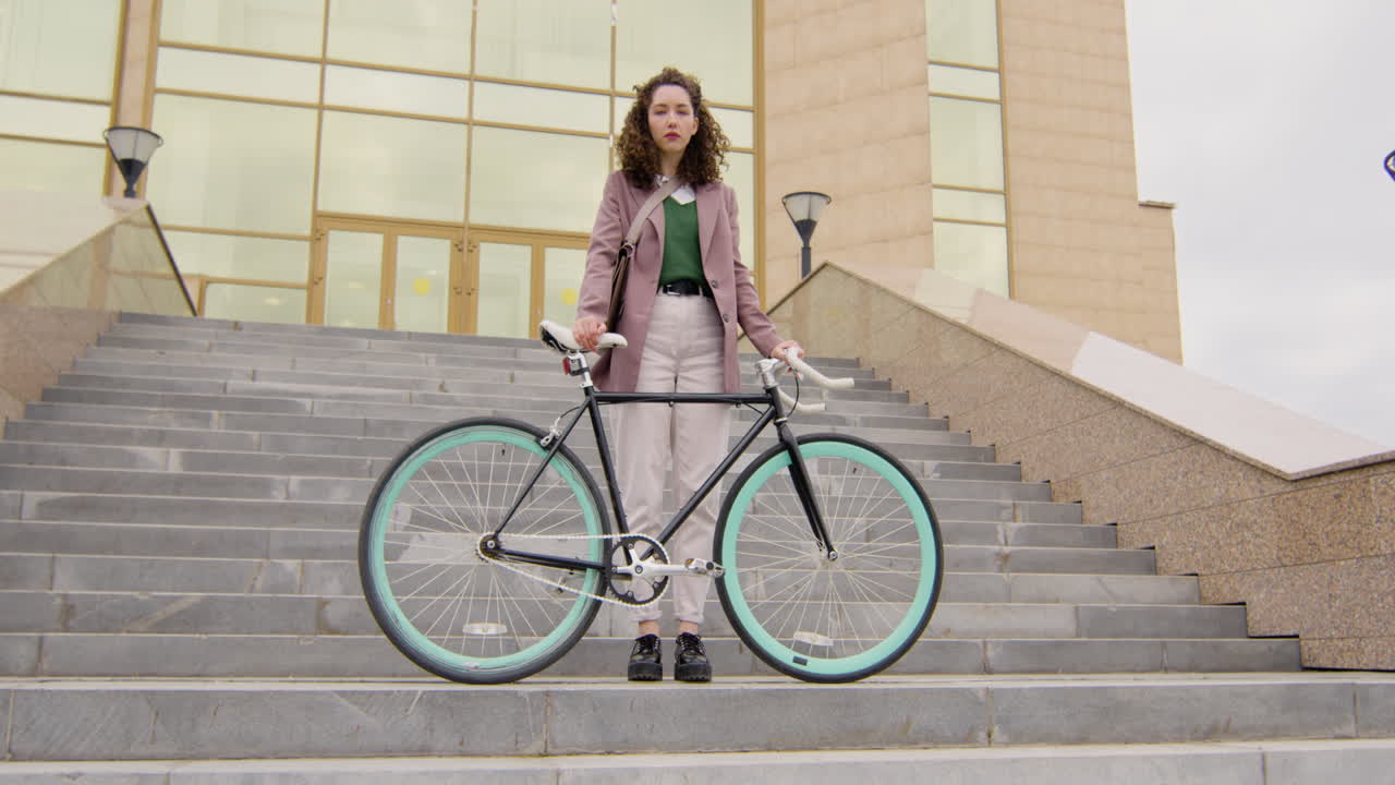 그녀의 자전거와 함께 계단에 서있는 동안 카메라를 찾고 정식 옷에 심각한 곱슬 여자
