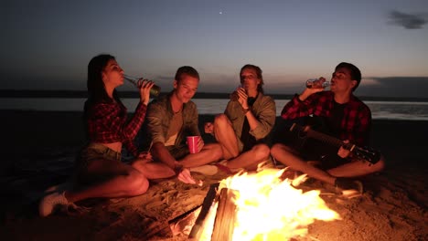 grupo de personas están pasando el tiempo cerca de la hoguera en la playa por la noche. bebiendo alcohol, saludos. joven está sosteniendo una guitarra. vista frontal