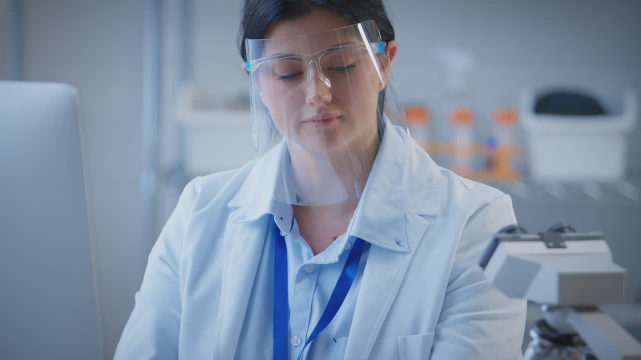ppe 얼굴 방패를 착용 한 여성 실험실 직원이 컴퓨터에 테스트 결과를 기록합니다.